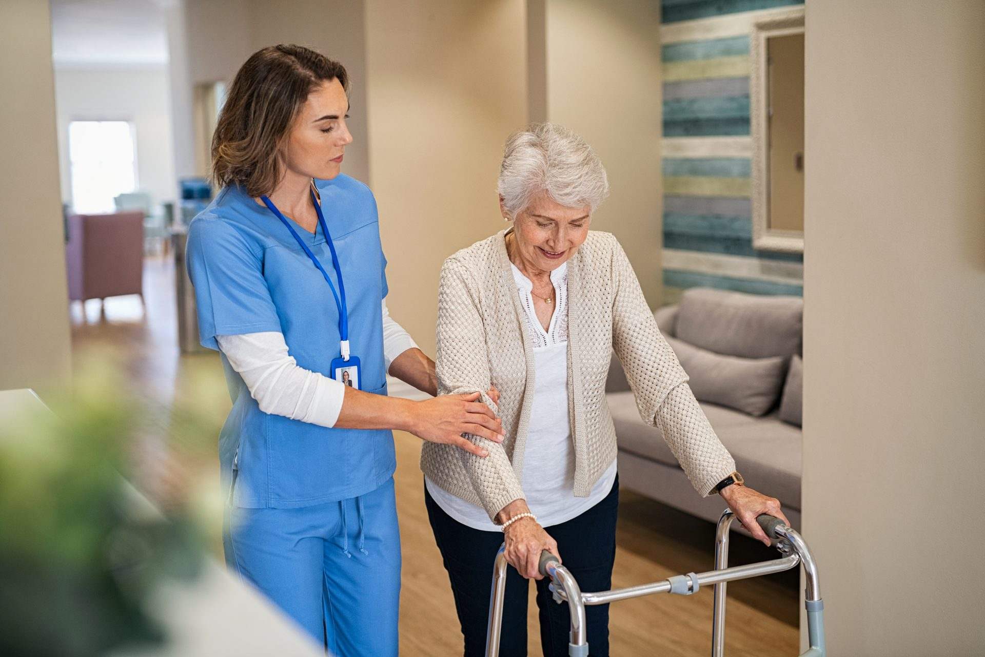 A carer in blue scrubs steadies a smiling older woman walking with a frame in a care home
