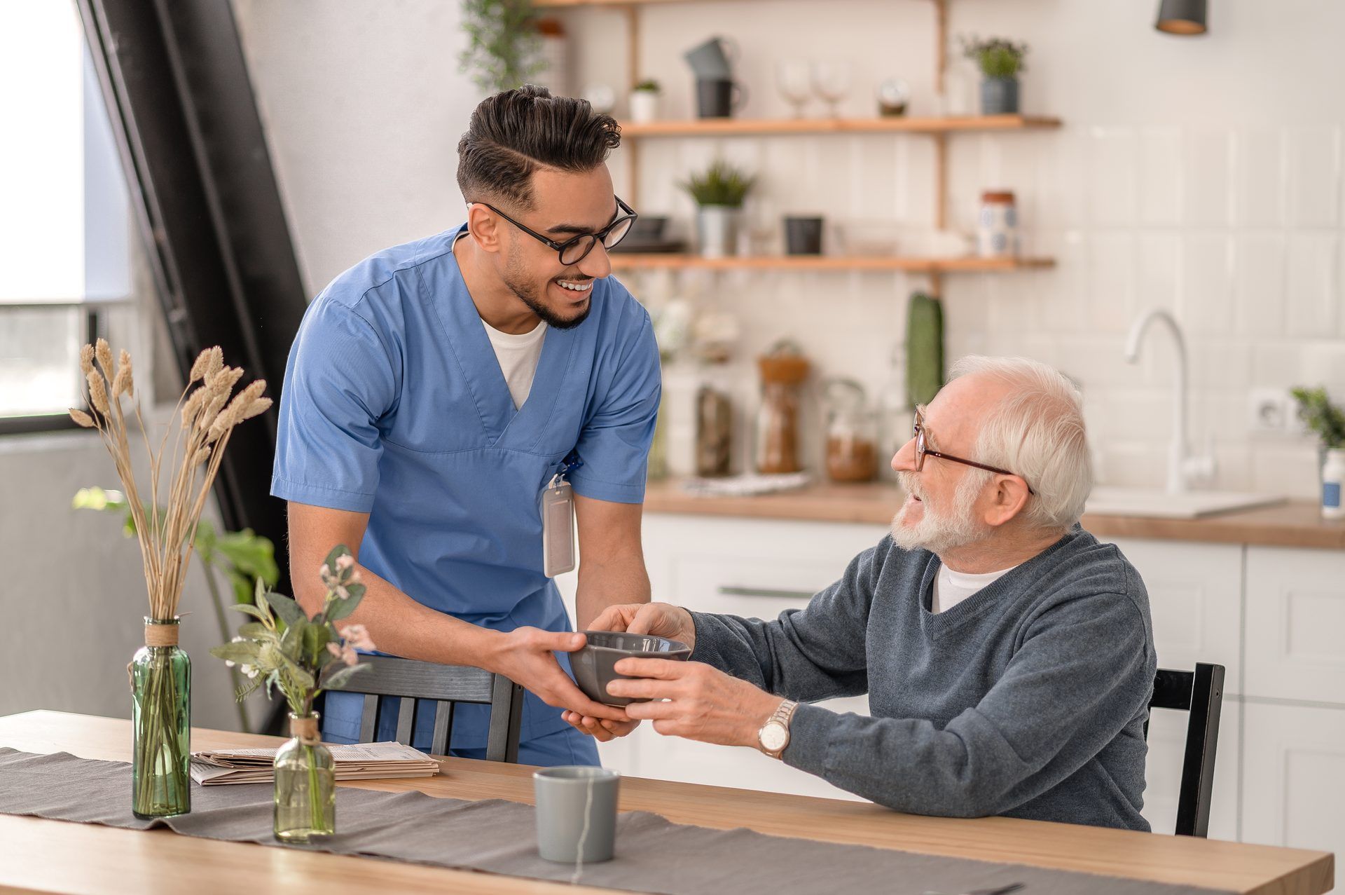 A care Professional in blue scrubs hands a bowl to a smiling older man at his kitchen table