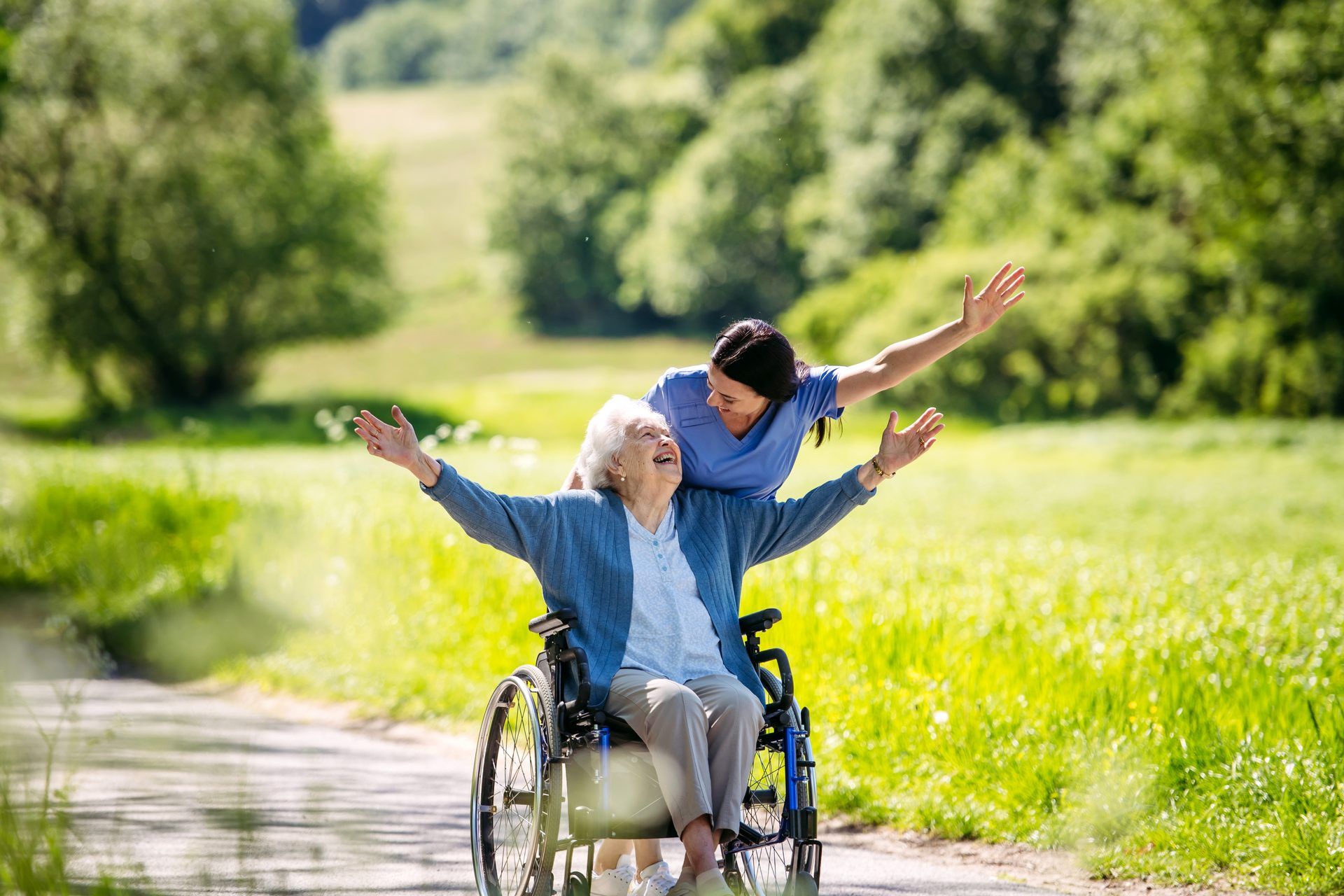 A carer in blue scrubs and a smiling older woman in a wheelchair stretch their arms out on a sunny country path